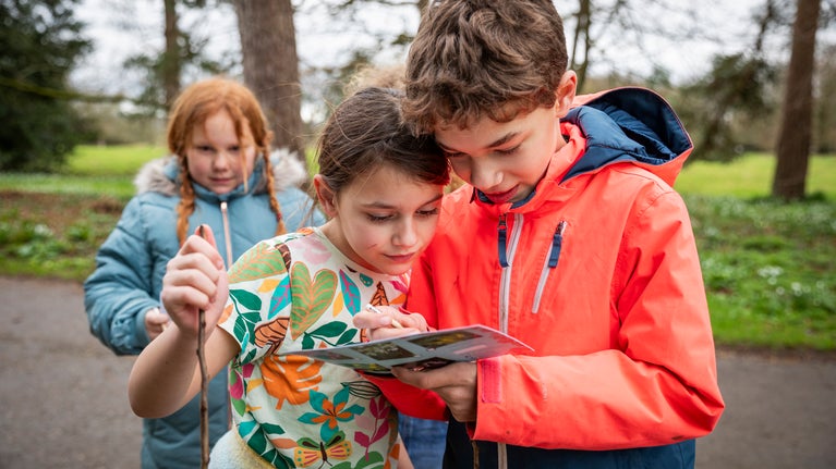 Two children looking closely at a trail sheet, with another child and greenery in the background.